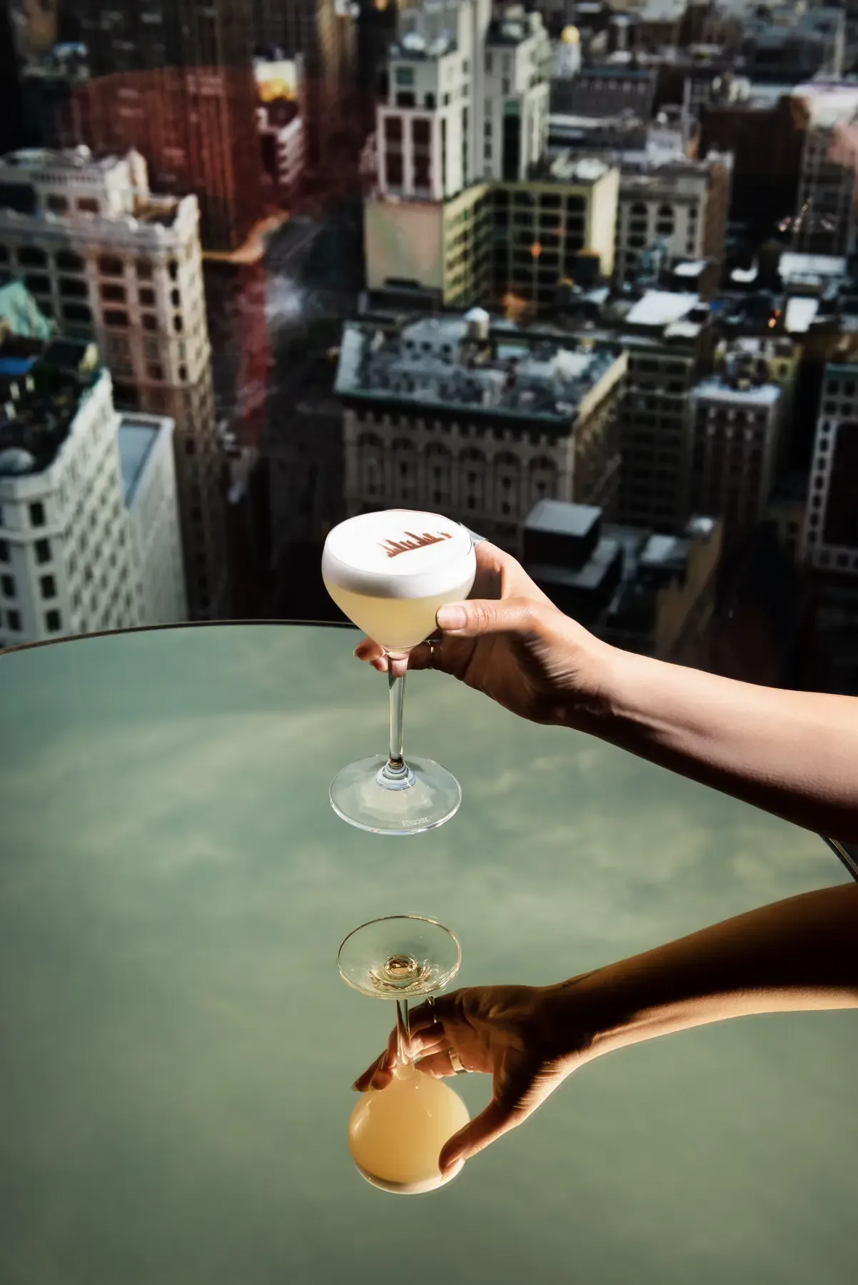 A foamy cocktail held above a reflective table with the Manhattan skyline beyond