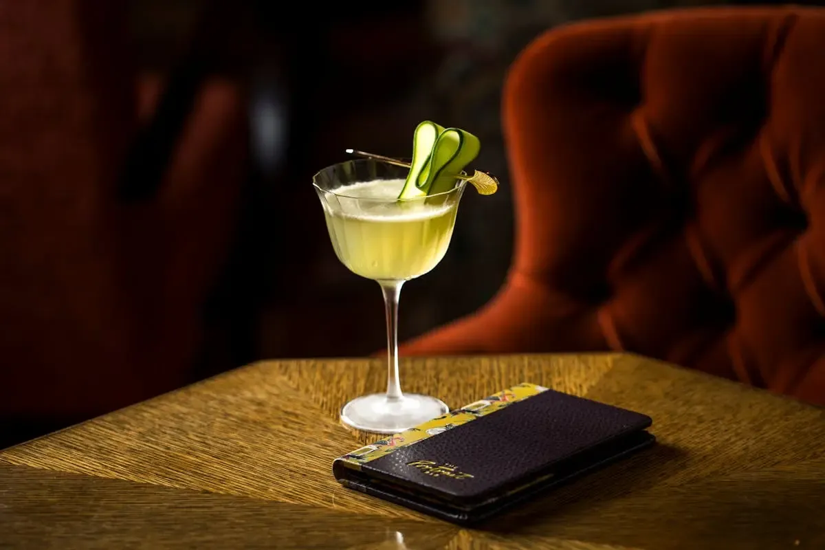 A pale green cocktail on a wood table beside a velvet banquette at The Portrait Bar