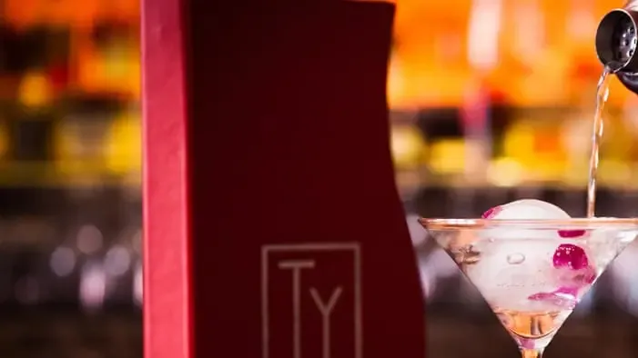 A bartender pouring a clear cocktail beside the red TY Bar menu